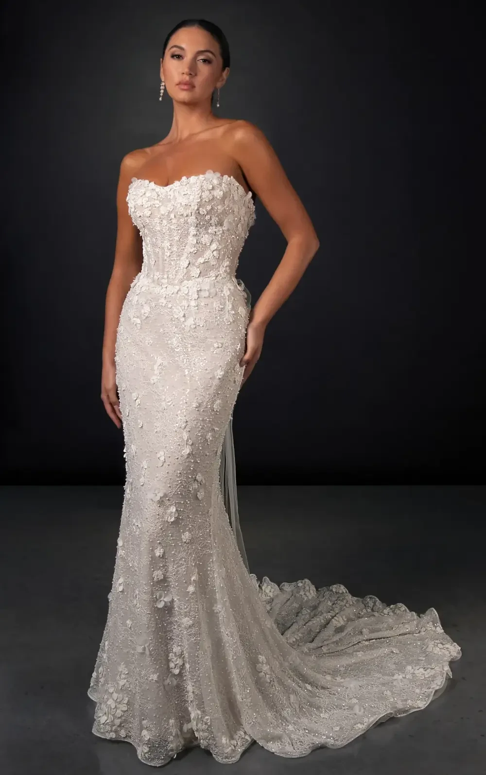 A woman wearing a strapless, fitted white gown with floral detailing, posing against a dark background.