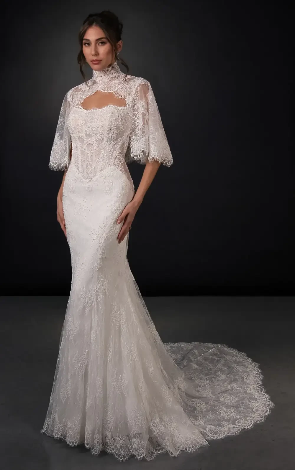 A model wearing a white lace wedding dress with a fitted bodice and a flowing train, posed against a dark background.
