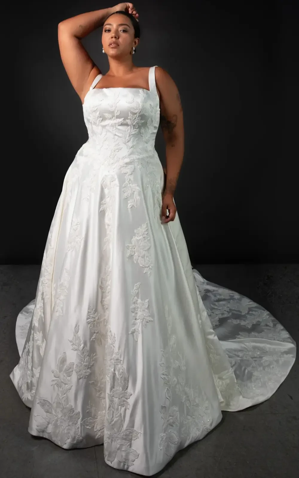 A model poses in a formal white wedding dress with intricate floral designs and a long train.